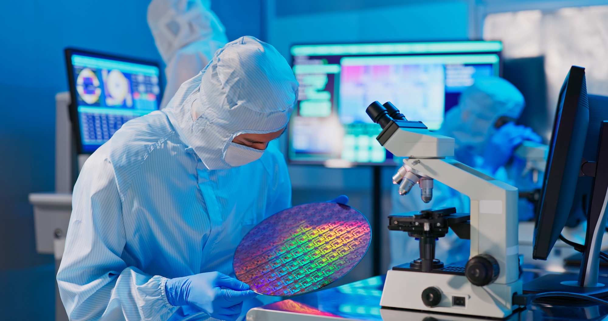 A male technician in a sterile coverall and gloves inspects a multicolored wafer at a semiconductor manufacturing plant, utilizing electrochemical deposition.