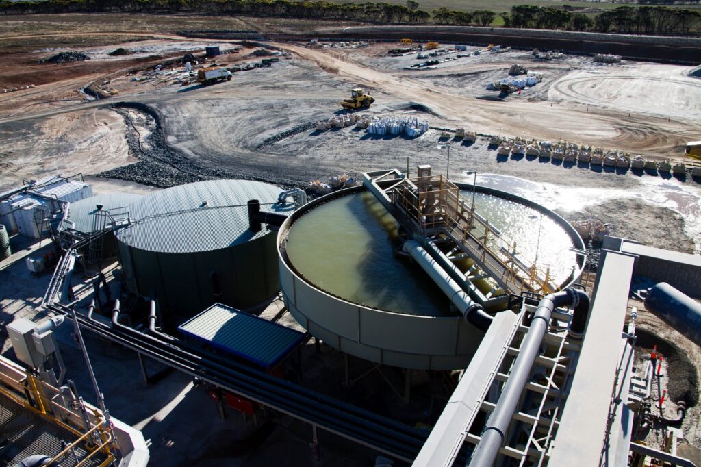 Aerial view of an industrial lithium processing facility with large circular tanks, pipelines, and machinery. The tanks are filled with liquid used in the separation or refining process. Surrounding the plant is a wide expanse of dirt and gravel, with construction equipment, vehicles, and storage areas visible in the background.