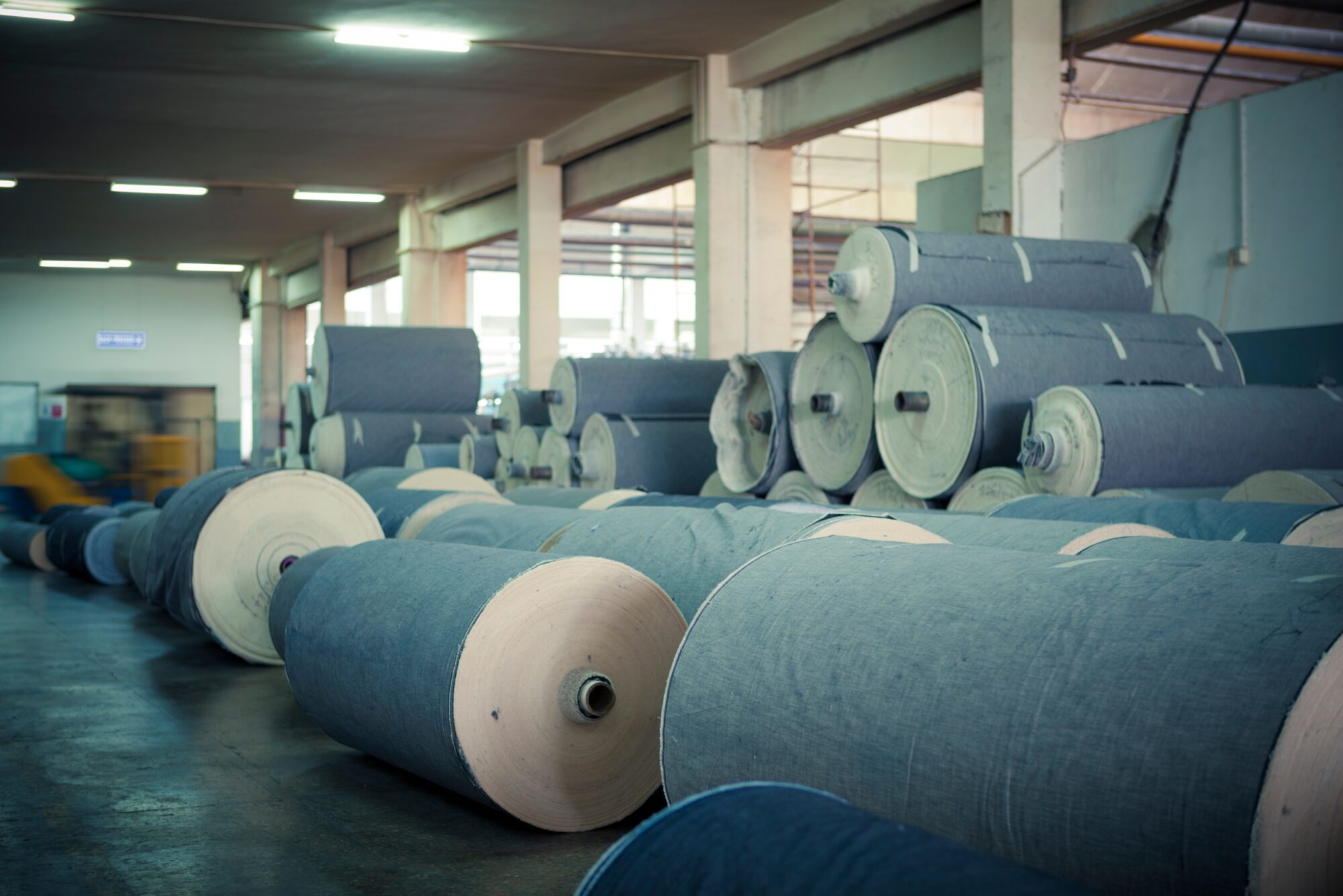 Large rolled denim fabrics stacked across a textile mill floor, illustrating industrial denim production and zinc dust finishing processes.