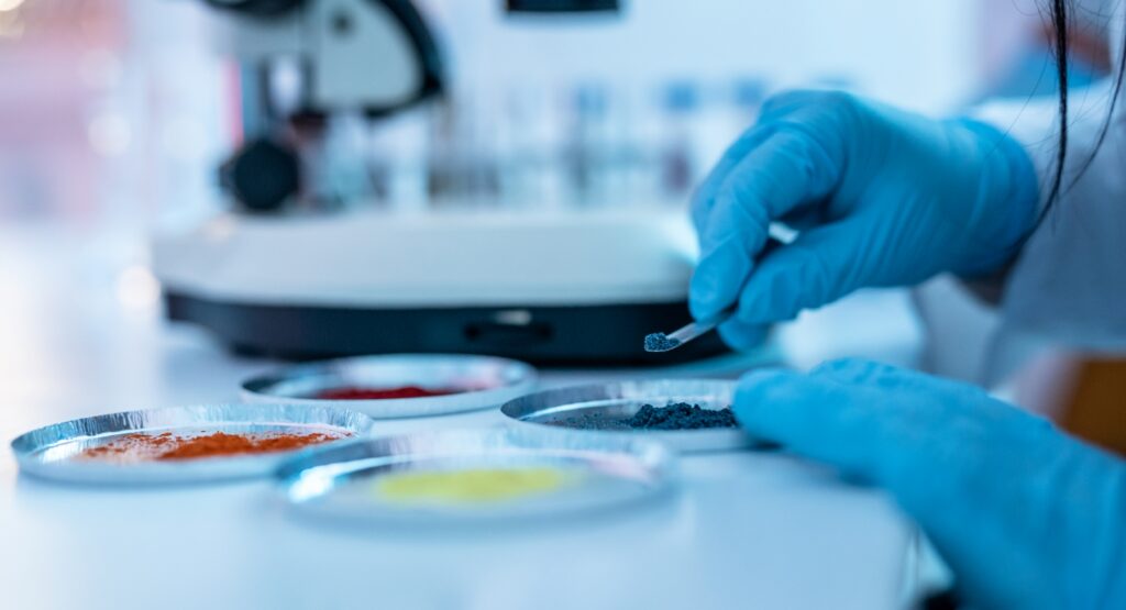 Laboratory researcher wearing blue protective gloves uses a metal spatula to examine a dark powdered material, with additional colored powder samples and laboratory equipment in the background.