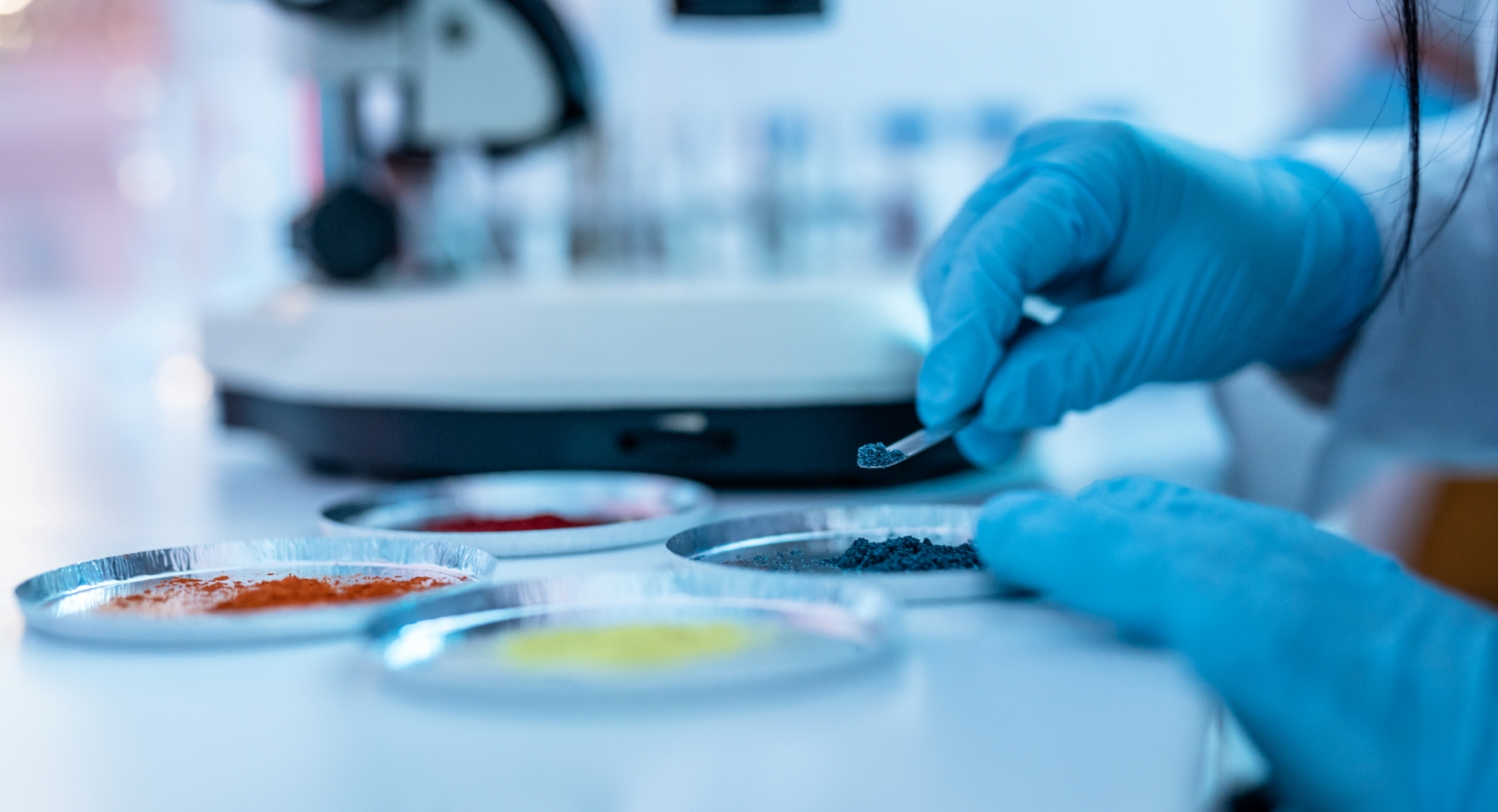 Laboratory researcher wearing blue protective gloves uses a metal spatula to examine a dark powdered material, with additional colored powder samples and laboratory equipment in the background.