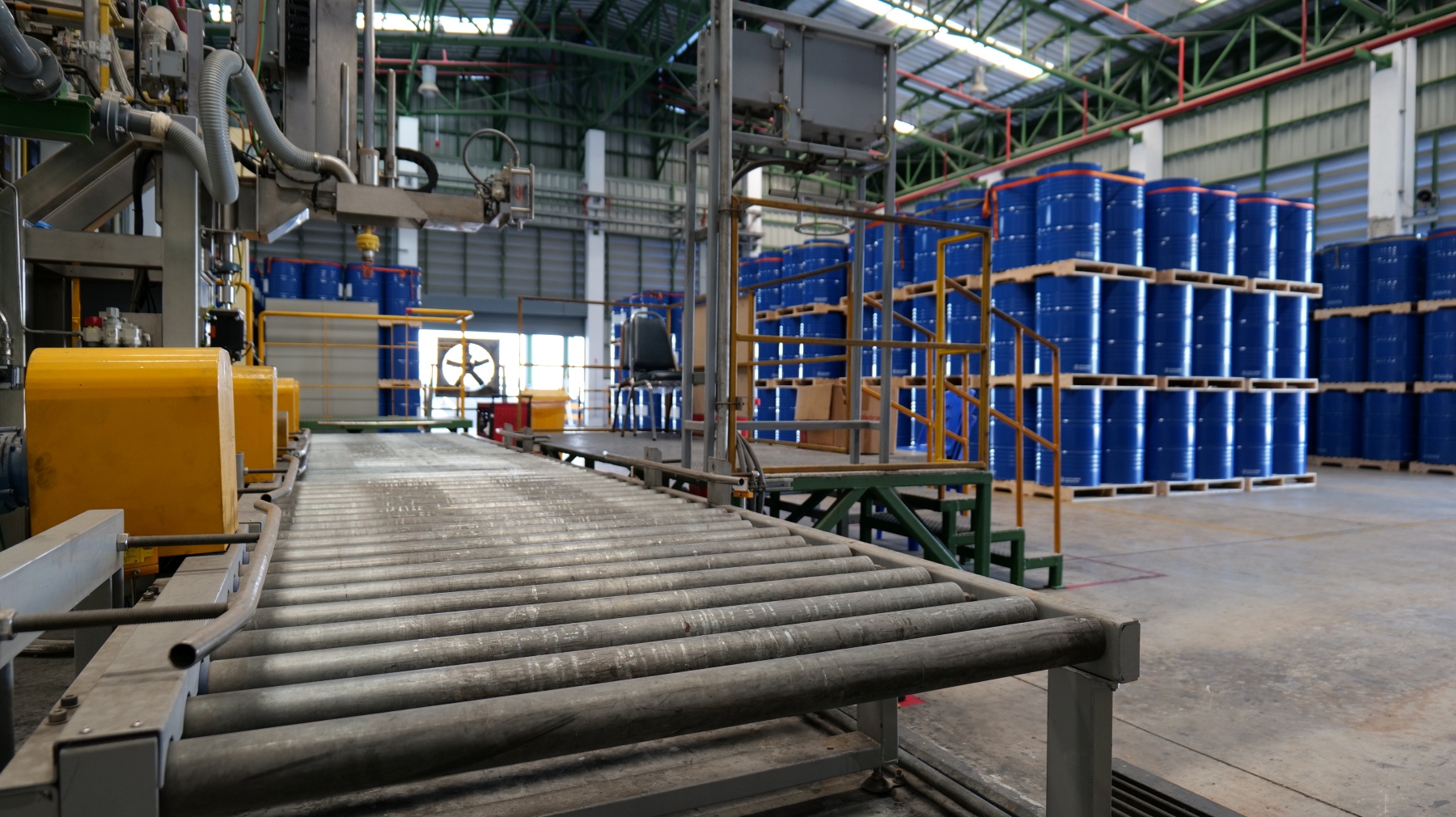 Industrial manufacturing facility featuring a metal roller conveyor system, automated processing equipment, and rows of blue storage drums stacked on wooden pallets.